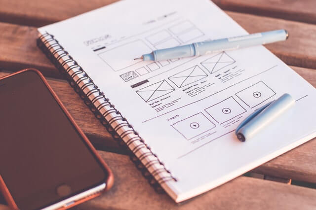 A notebook with a website layout sketched on its page sitting next to a phone and pen on a wooden table.